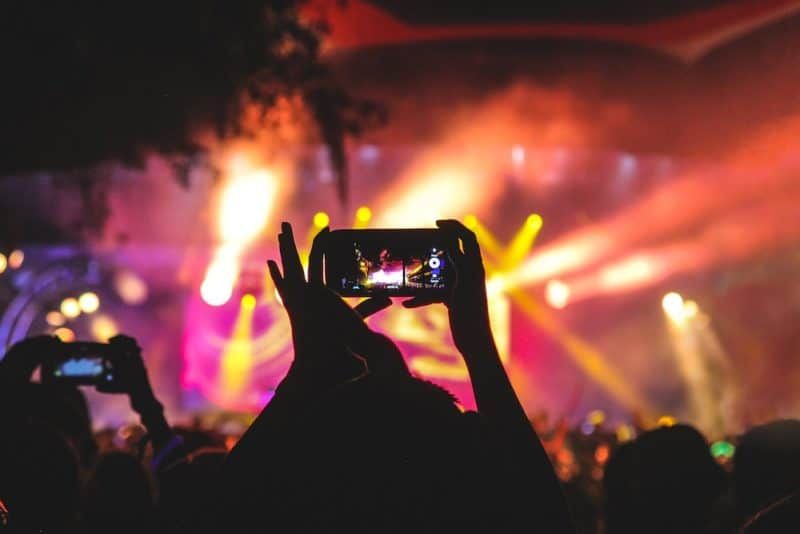 Persona que tiene un teléfono inteligente tomando un video de un concierto cerca del escenario con luces durante la noche